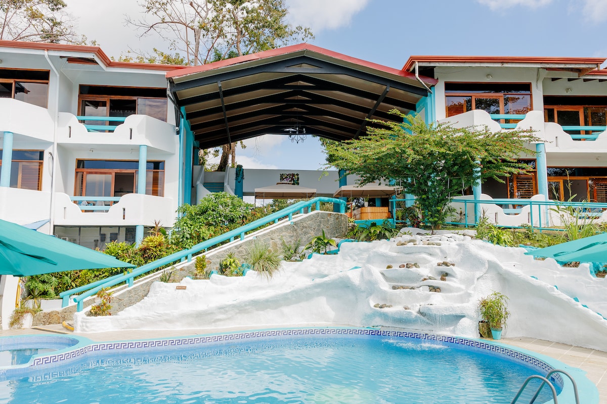 An inviting pool area is framed by a gently sloping hillside, with cascading steps leading to the entrance of the hotel. The structure features a distinctive overhanging roof and is surrounded by lush greenery, creating a serene environment for relaxation.