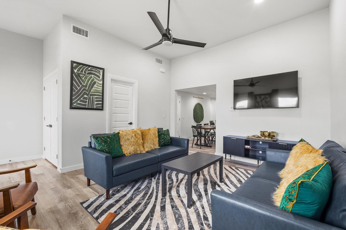 The living area features two navy sofas adorned with decorative pillows and a cozy throw. A black coffee table sits centrally on a striped area rug, while a wall-mounted television is visible across the room. Natural light filters through the open space, enhancing the modern decor.