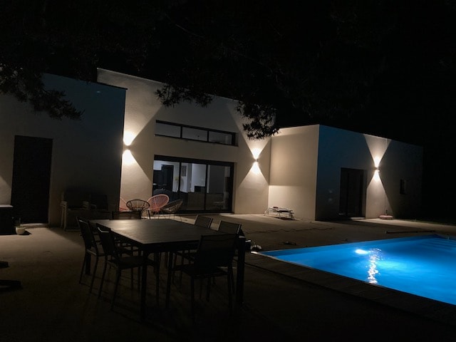 The modern exterior of a house is shown at night, featuring recessed lighting that highlights the architectural lines. The illuminated swimming pool reflects a gentle blue hue, while a dining table with chairs sits on the patio, offering a setting for outdoor gatherings.