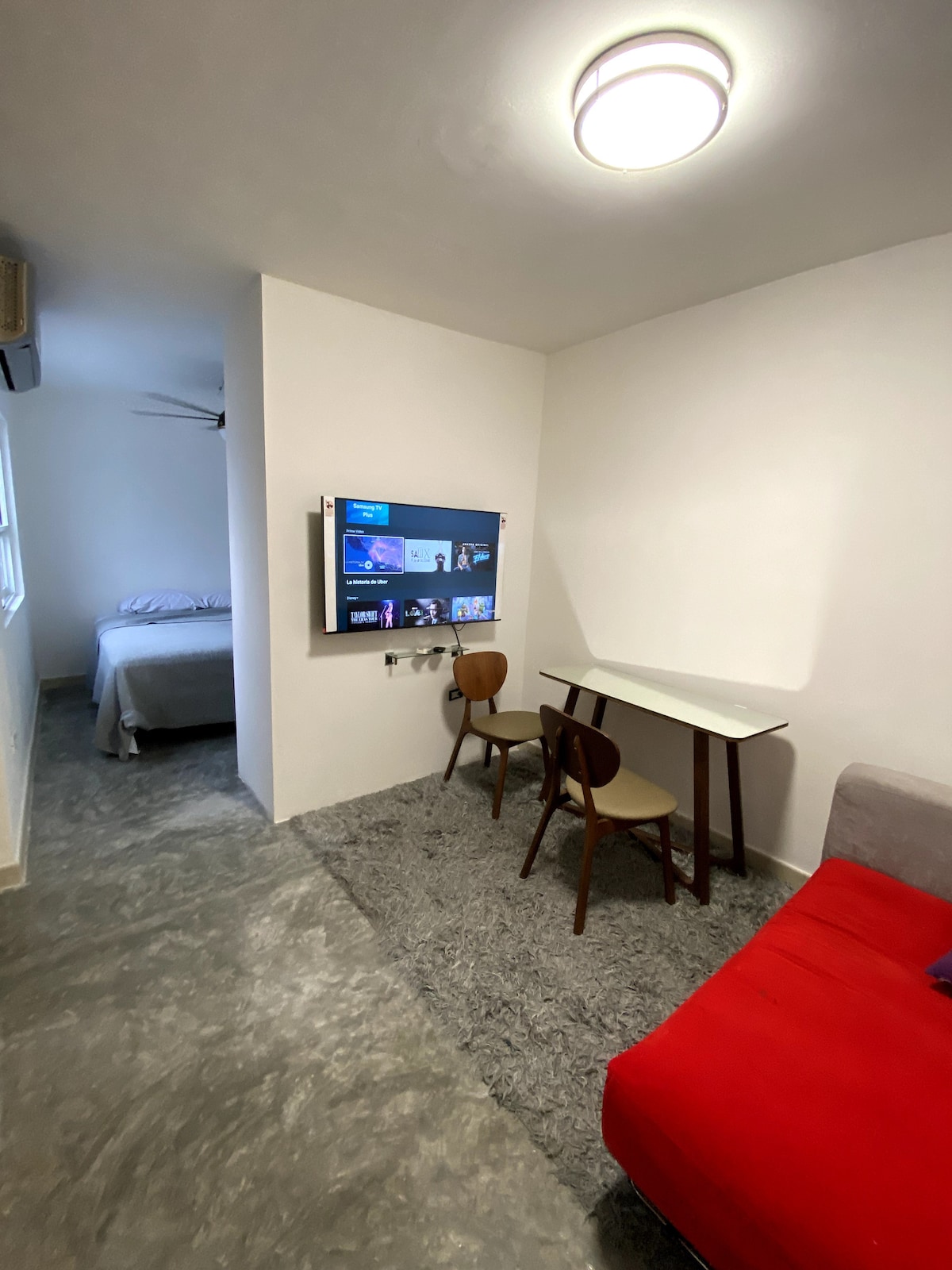 A compact living space features a red sofa in the foreground and a small dining table with two chairs. A wall-mounted television displays content across from a sleeping area, which includes a spacious bed in the background, and light comes through a nearby window.
