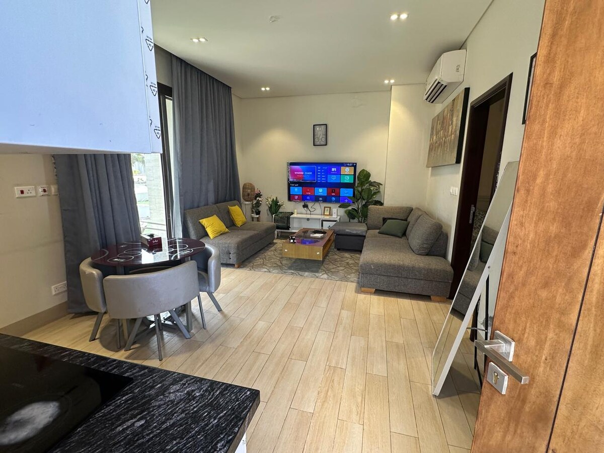 A modern living space features a comfortable sectional sofa and armchairs arranged around a low coffee table. A large television is mounted on the wall, surrounded by soft lighting and decorative elements. Large windows allow natural light to brighten the room.