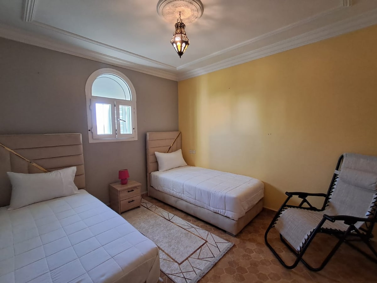 A tranquil sleeping area features two spacious beds with light bedding. A nightstand with a pink lamp is positioned between them. A folding chair adds seating. Natural light enters through a window with a decorative arch above, while the walls are painted in soft grey and warm yellow tones.