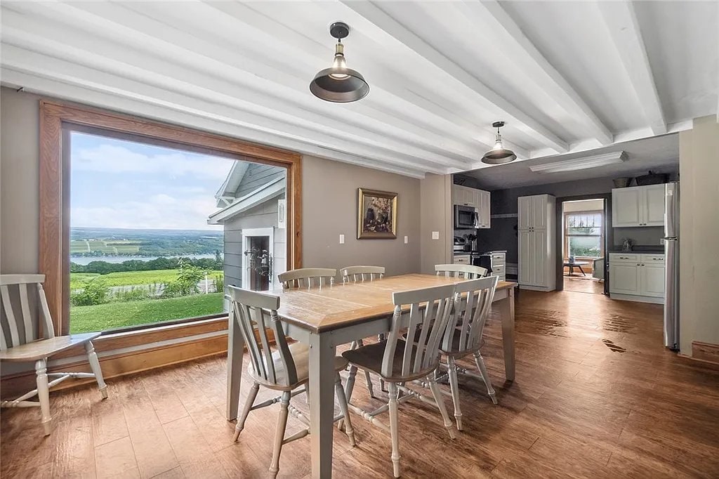 A dining area features a large wooden table surrounded by six light-colored chairs. A wide window offers expansive views of the surrounding landscape and lake, while the space is illuminated by overhead pendant lights. The kitchen is partially visible in the background.