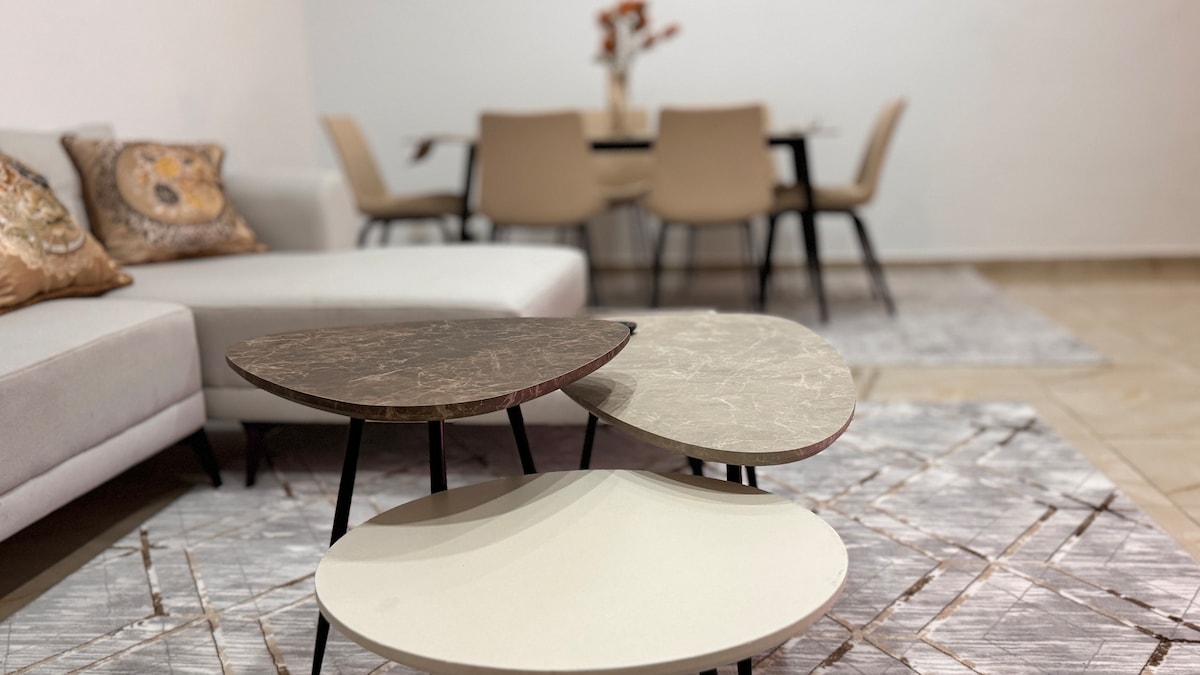A modern living area features a light-colored, spacious sofa adorned with decorative pillows. Two round coffee tables with various surface finishes are positioned in front of the sofa. A dining table with chairs is visible in the background, complemented by a textured area rug.