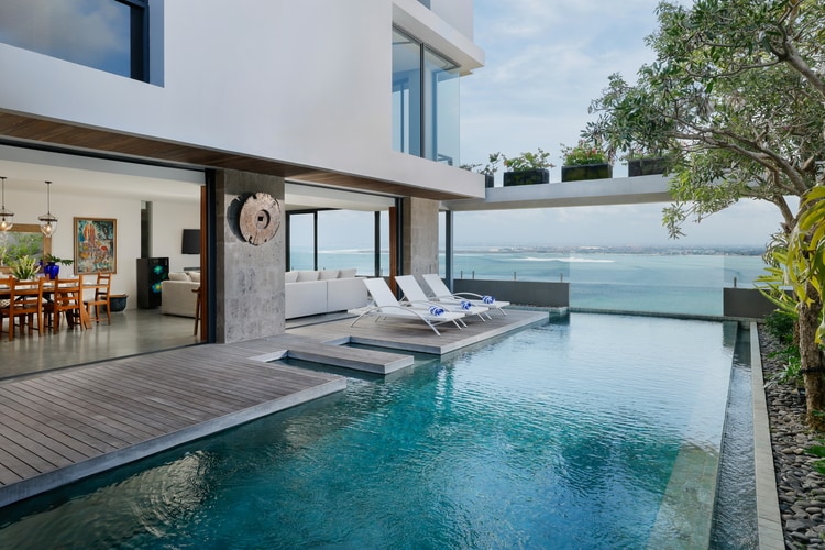 An outdoor area features a private pool reflecting the blue sky and surrounded by a wooden deck. Sun loungers are positioned beside the pool, overlooking expansive sea views. The adjacent living area includes a dining table set for six, enhancing the indoor-outdoor living experience.