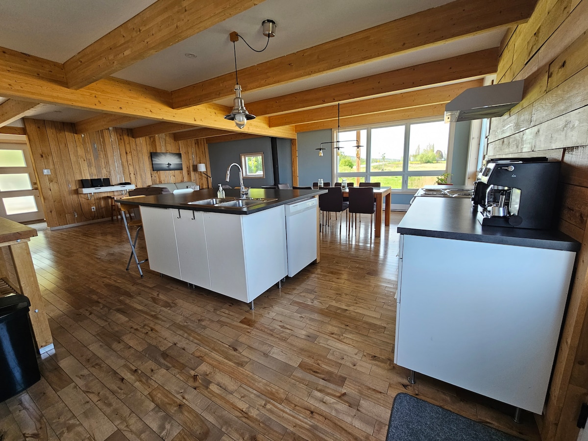 A spacious kitchen and dining area feature natural wood beams and ample lighting from large windows. A central island with seating is complemented by modern appliances and sleek cabinetry, creating a functional space for cooking and dining with views of the surrounding landscape.