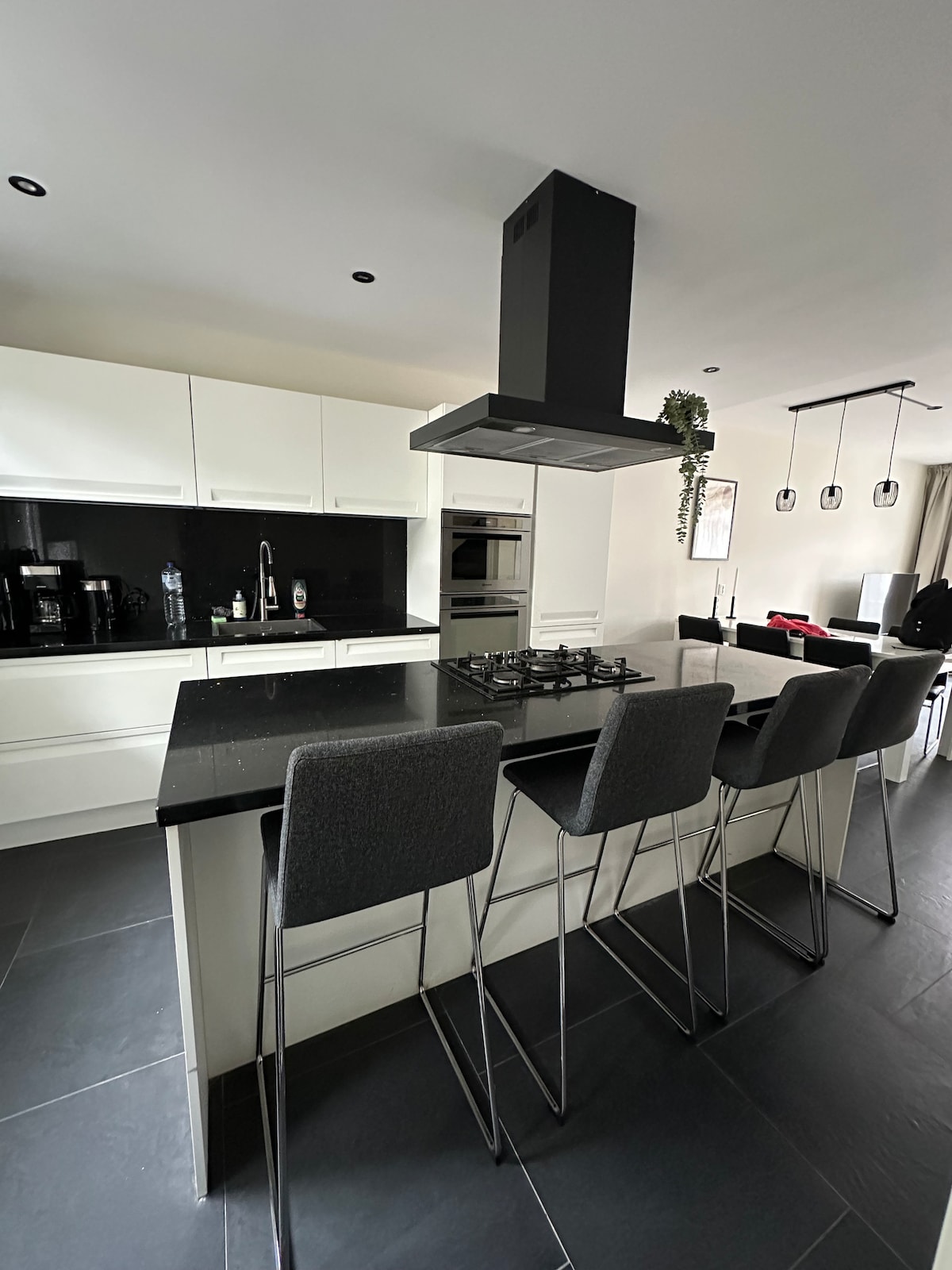 A modern kitchen is highlighted by sleek white cabinetry and a large black cooking island with bar stools. A stainless steel exhaust hood is present above the cooking area, while built-in appliances like an oven and refrigerator are integrated into the design. Natural light enters through nearby windows.