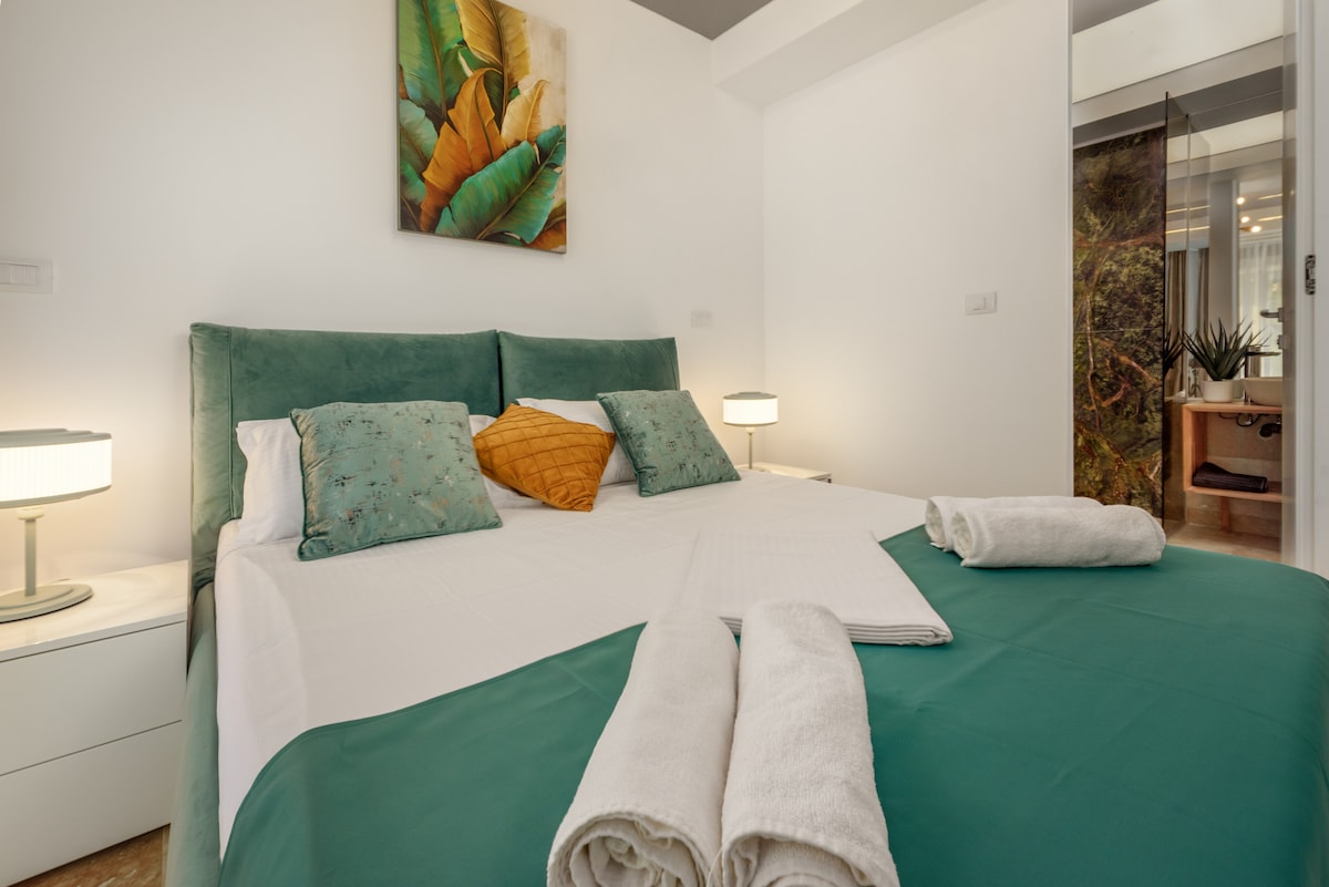 A spacious sleeping area features a large, upholstered bed with green and white bedding. Two contrasting pillows add texture, while lamps on either side provide soft illumination. A colorful wall art piece depicting tropical leaves enhances the calming ambiance.