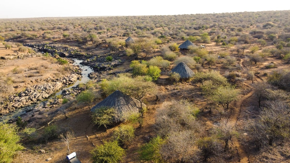 An aerial view captures the tranquility of the landscape, showcasing traditional bandas with thatched roofs nestled within a natural setting. The Ewaso Nyiro river meanders through the dry terrain, surrounded by sparse vegetation and rocky outcrops, offering a sense of seclusion and connection to nature.