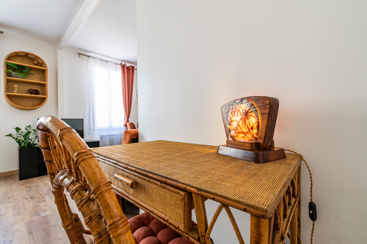 A rattan desk is positioned near a window, creating a bright workspace. An artisan lamp sits on the desk, featuring a warm glow with a decorative design. A comfortable chair complements the desk, while a plant is visible in the background.