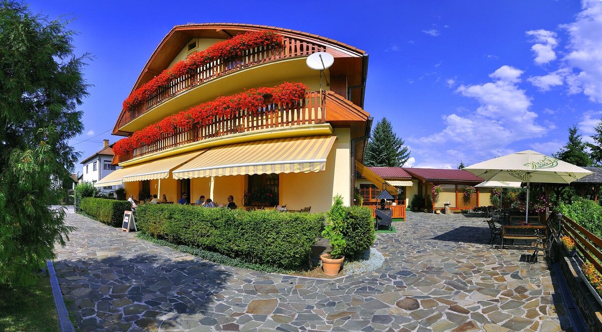 A three-story building features colorful balconies adorned with flowering plants. A patio area is visible in front, with shaded seating beneath an umbrella. Stone pathways lead through well-maintained greenery, complemented by a clear blue sky with scattered clouds.