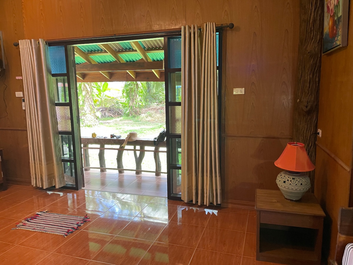 An open space reveals glass doors leading to an outdoor sitting area, framed by light beige curtains. A decorative lamp rests on a wooden side table, and a small woven mat lies on the polished floor. Lush greenery is visible through the doors.
