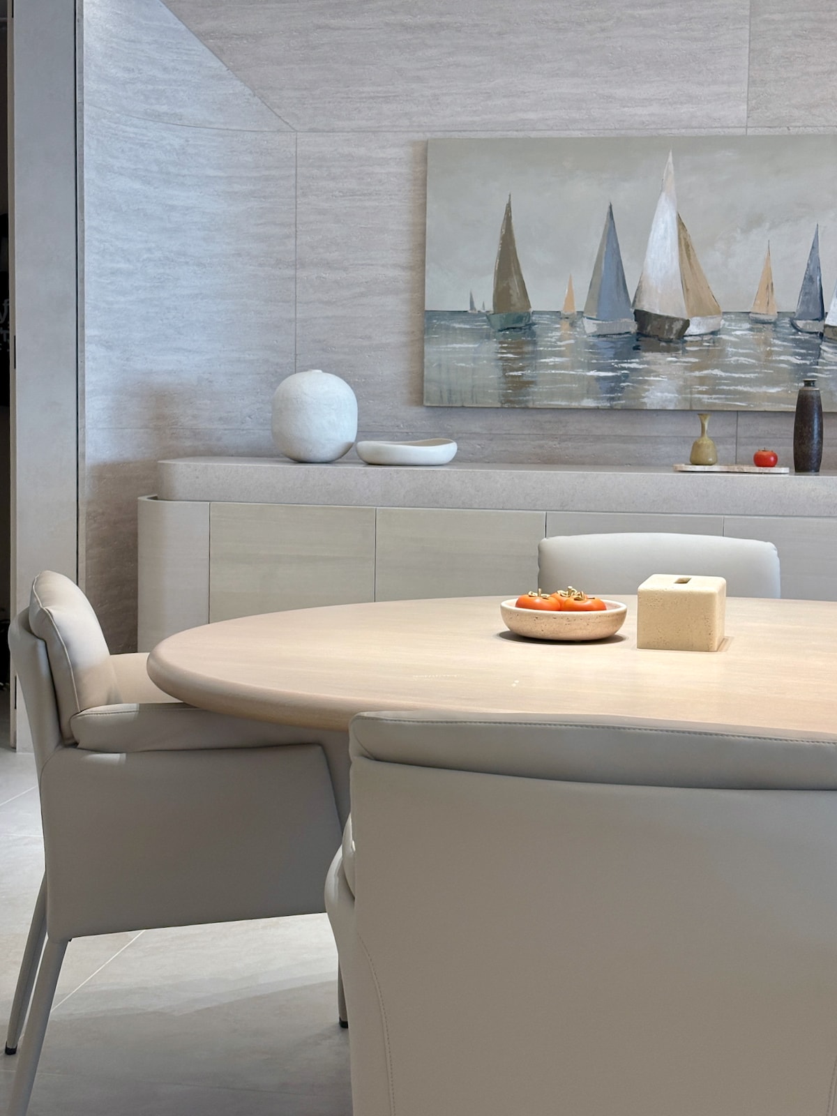 A modern dining area features a round wooden table surrounded by stylish cream-colored chairs. A serene painting of sailboats is displayed on the wall, while a centerpiece bowl holds fresh oranges. Natural light enhances the soft tones of the space.