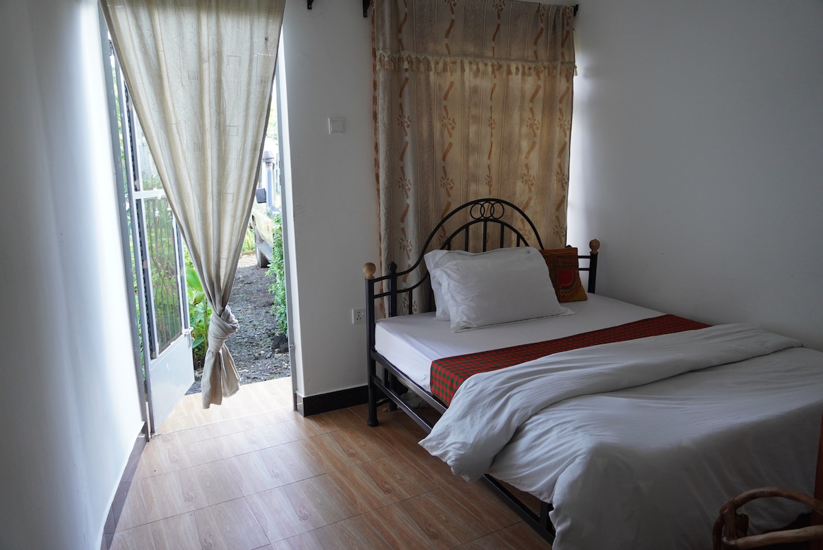 A cozy room features a spacious bed with a white comforter. Natural light floods in through a glass door leading to a garden area, while soft curtains frame the entrance. The warm wooden flooring complements the neutral wall color, creating a calm environment.