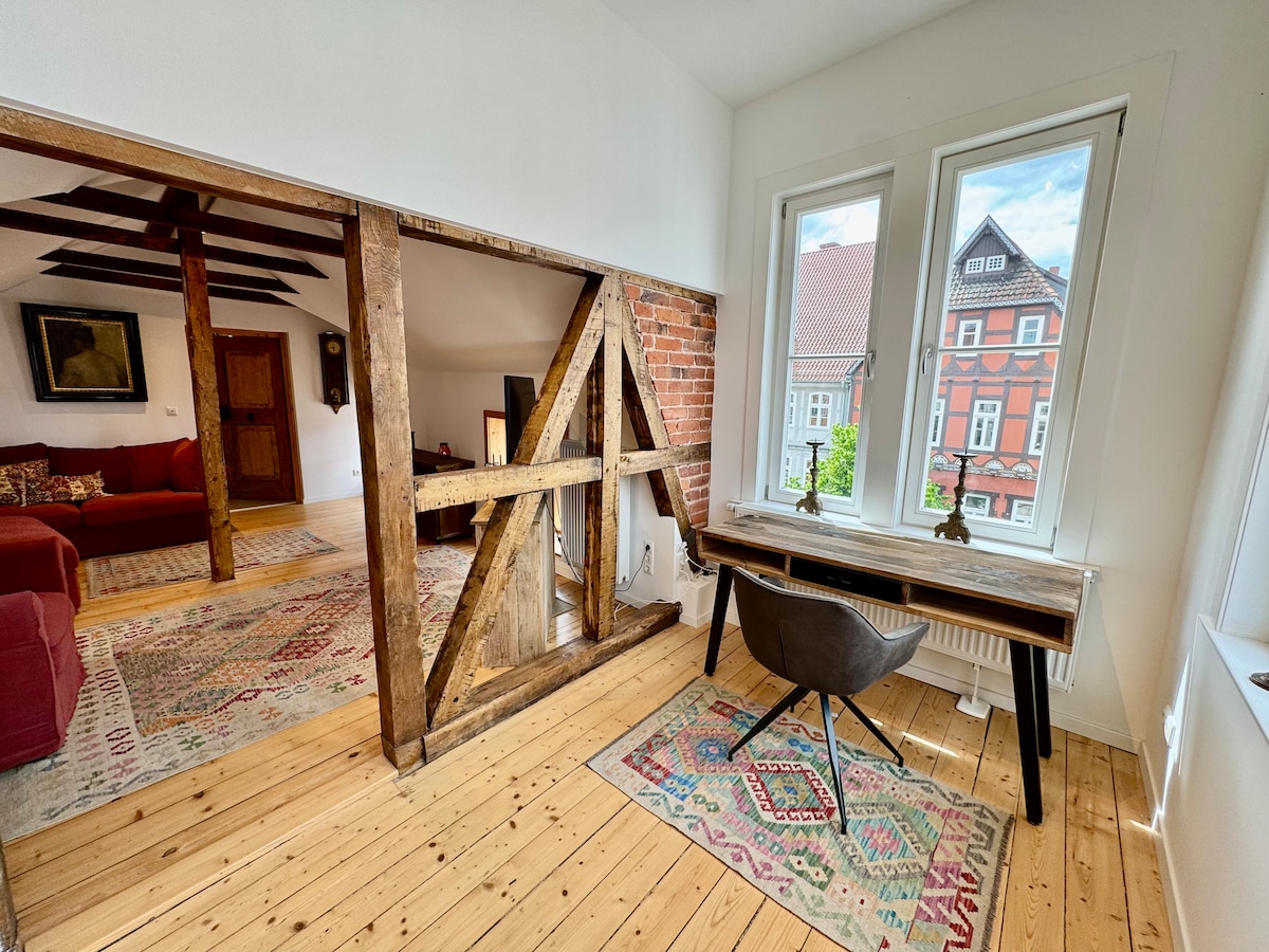 A workspace is situated near a large window, allowing natural light to illuminate the area. Rustic wooden beams frame the entryway to the adjacent living space. A patterned rug covers the wooden floor, and a stylish chair complements the wooden desk.
