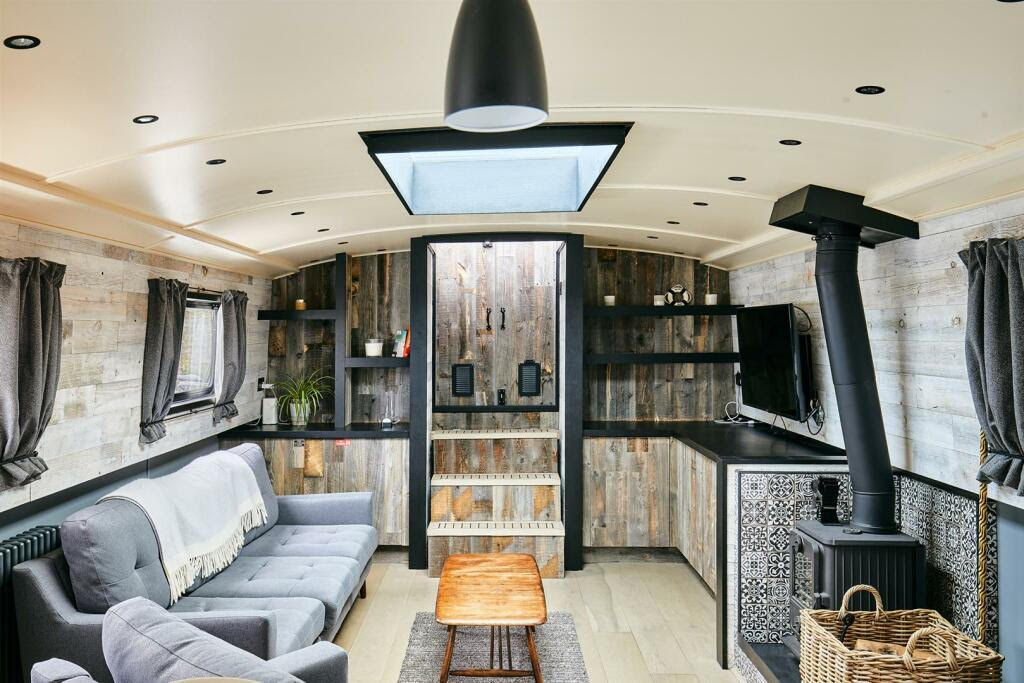 The interior of the houseboat features a cozy living area with a comfortable gray sofa and a wooden coffee table. Natural wooden accents are present on the walls, complemented by a log burner and a wall-mounted television. Large windows allow for ample light.