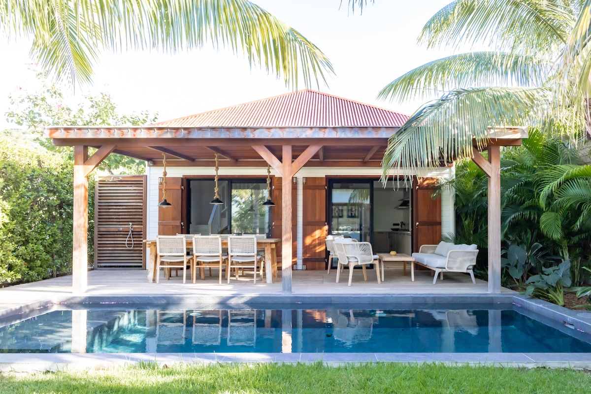 A charming wooden terrace is situated by a tranquil swimming pool, surrounded by lush greenery. Outdoor seating arrangements are present, along with a dining table. Large windows allow natural light to fill the interior space, enhancing the connection to the tropical environment.