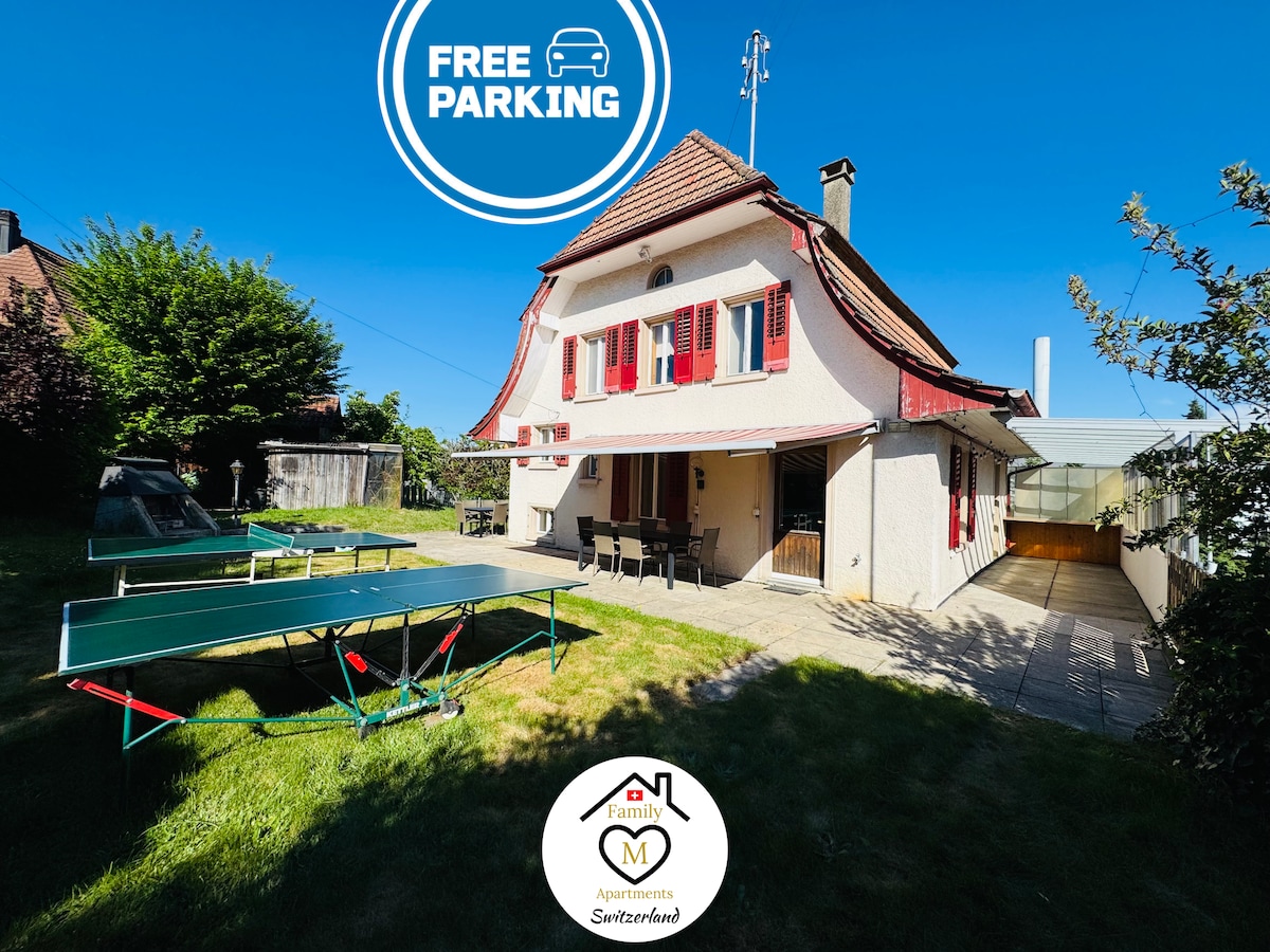 An outdoor area features dual table tennis tables positioned on green grass. Behind, a spacious house with a charming red-roofed design is visible. A patio with seating provides an inviting spot for relaxation. Clear skies and sunlight enhance the overall openness of the space.