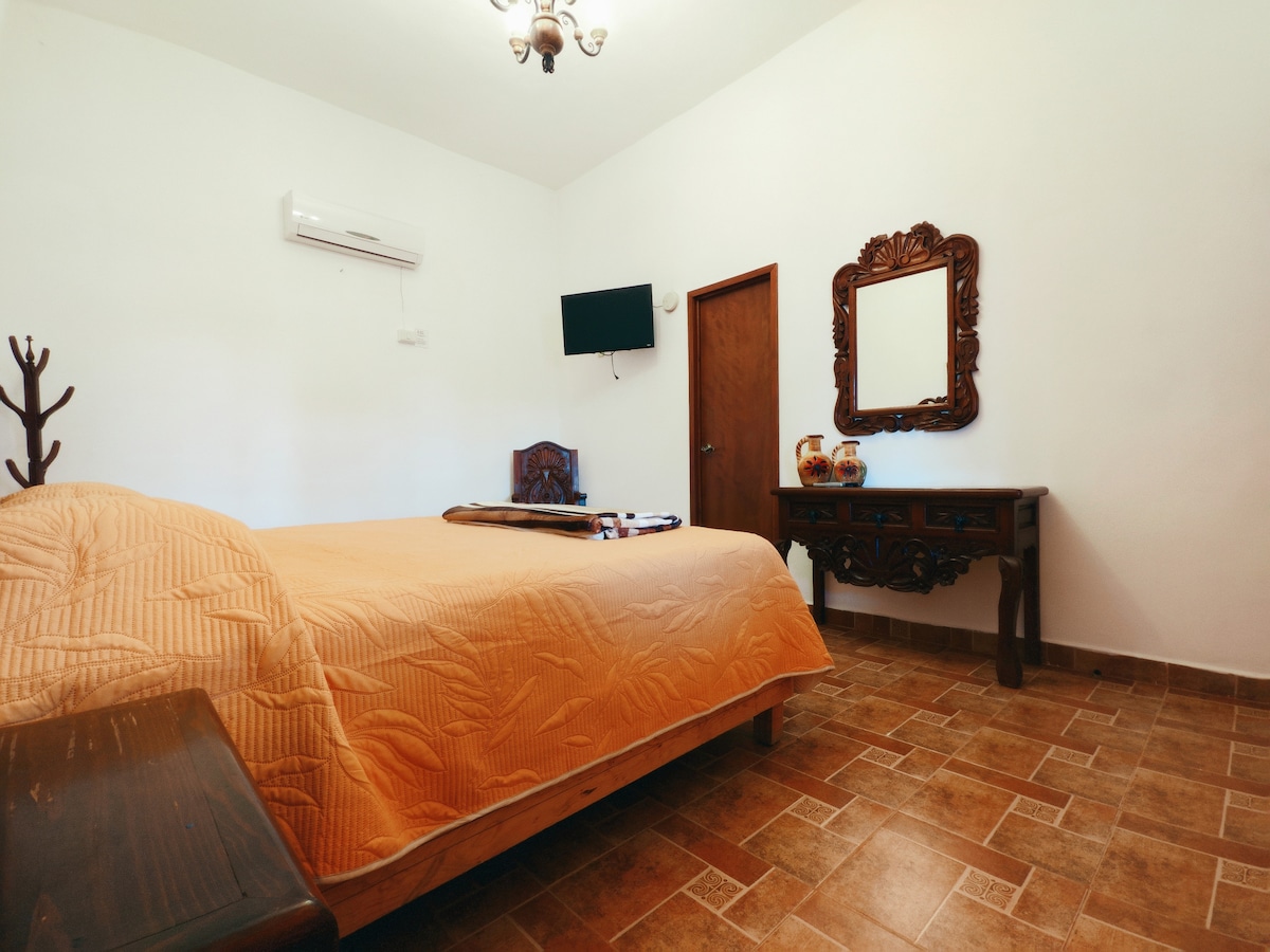 A well-lit bedroom features a spacious bed covered with an orange quilt. A decorative wooden mirror is mounted on the wall above a dark wood console table. Natural light enters through a window, and a flat-screen TV is mounted near the ceiling.