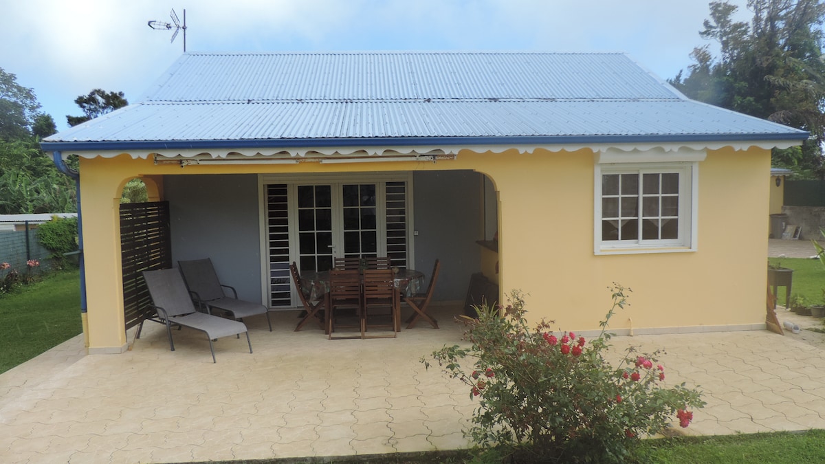 A charming exterior of a creole-style house is presented, featuring a spacious covered patio. Wooden furnishings, including a dining table and lounge chairs, are arranged for outdoor enjoyment. Lush greenery and a blooming rose bush add a touch of nature to the scene, while light clouds linger overhead.