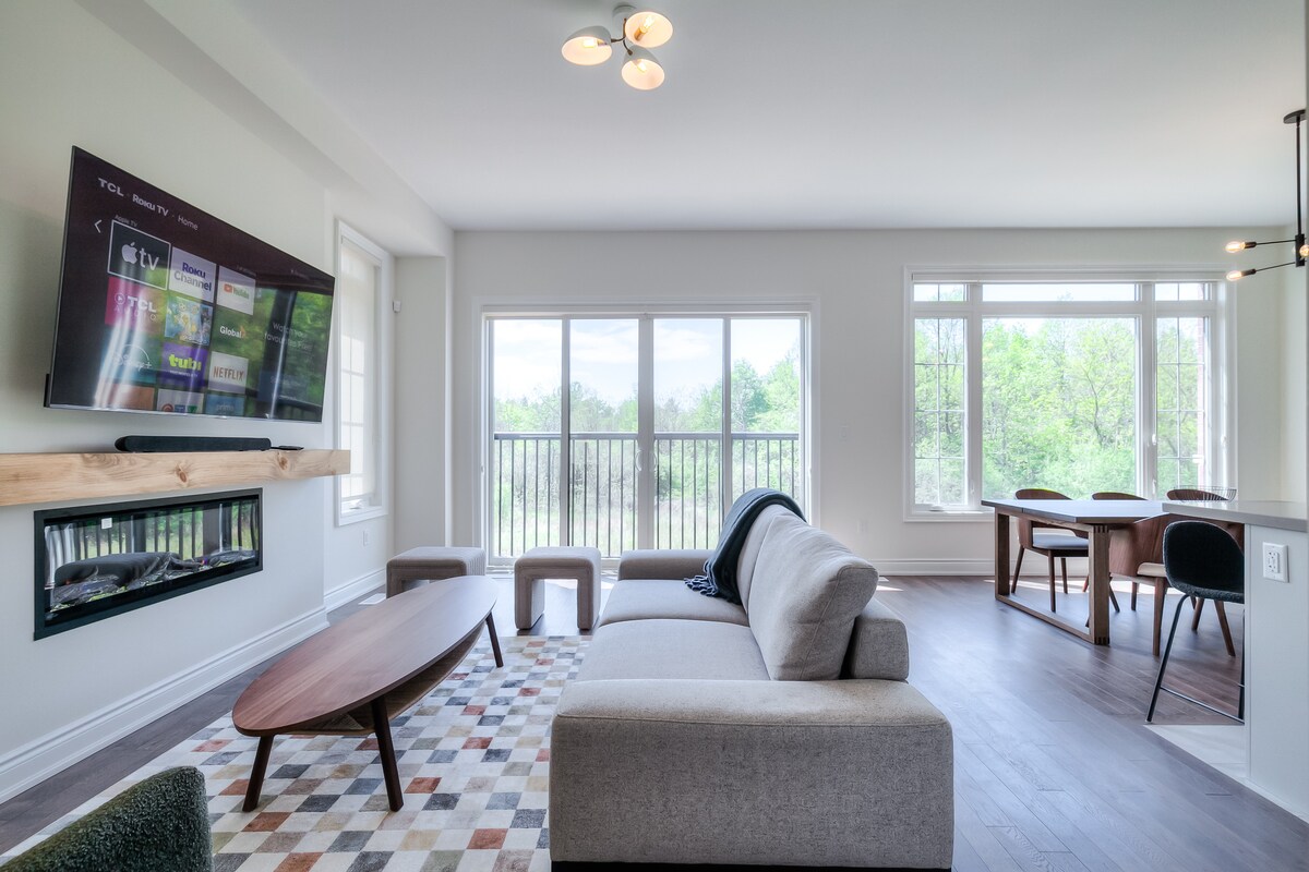 An open-concept living area is depicted with large windows allowing natural light to fill the space. A mounted TV sits above a sleek gas fireplace, and a light-colored sofa with accent pillows is positioned on a patterned area rug. A dining table with chairs is visible in the background.