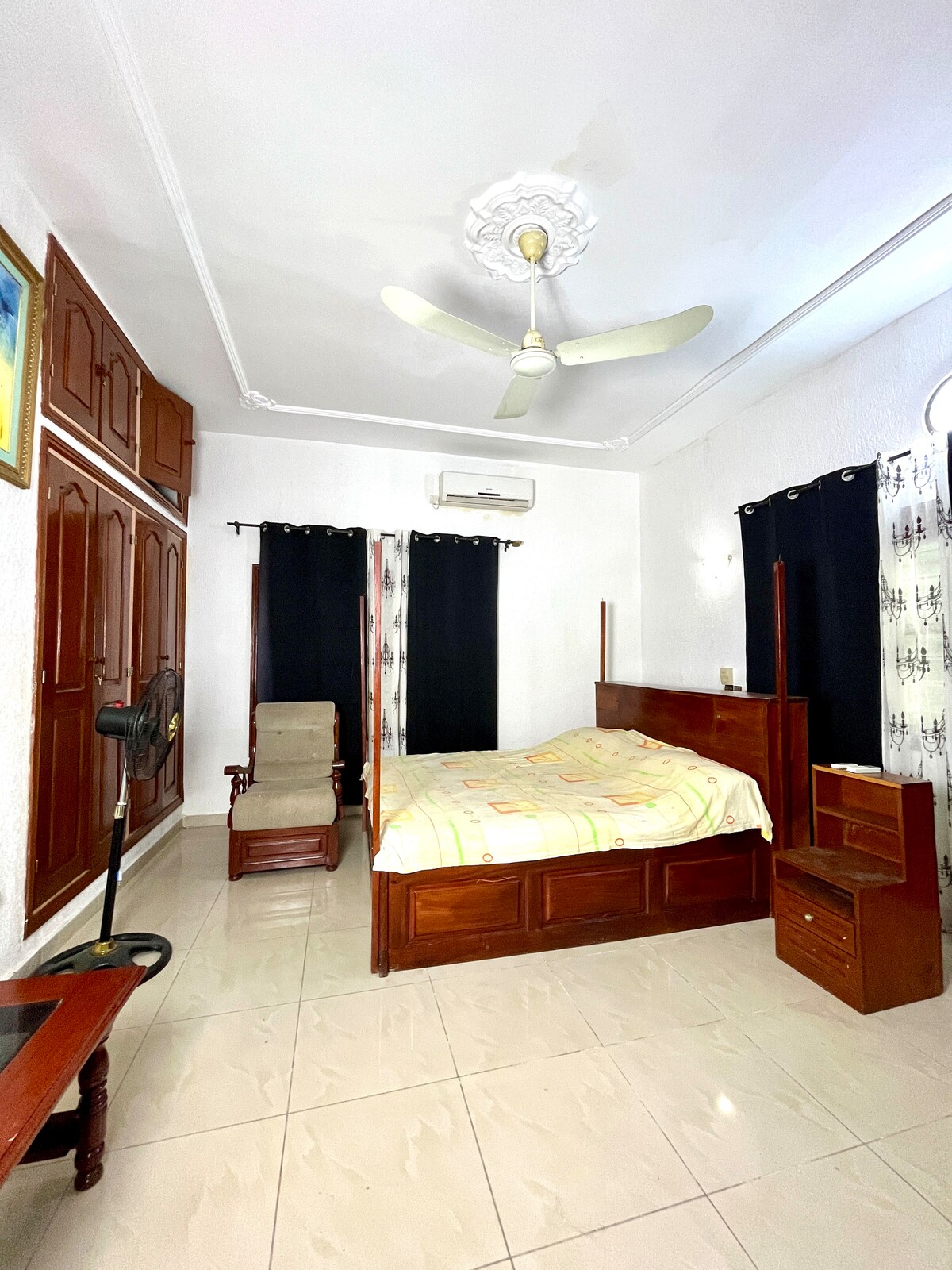A well-lit bedroom features a spacious bed with wooden furnishings and matching nightstands. Dark curtains provide privacy, while a ceiling fan is present for comfort. A wooden dresser and a seating area with an armchair are visible, enhancing the room's functionality.
