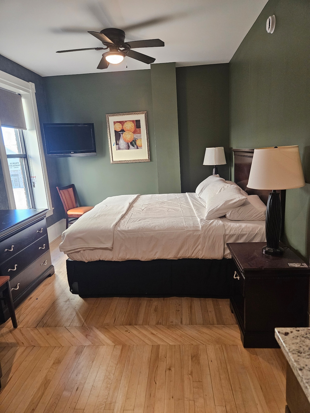 A sleeping area features a spacious bed with crisp white linens, complemented by dark wood furnishings. A flat-screen TV is mounted on the wall, while soft ambient lighting is provided by two lamps. A window allows natural light to bring warmth to the room.