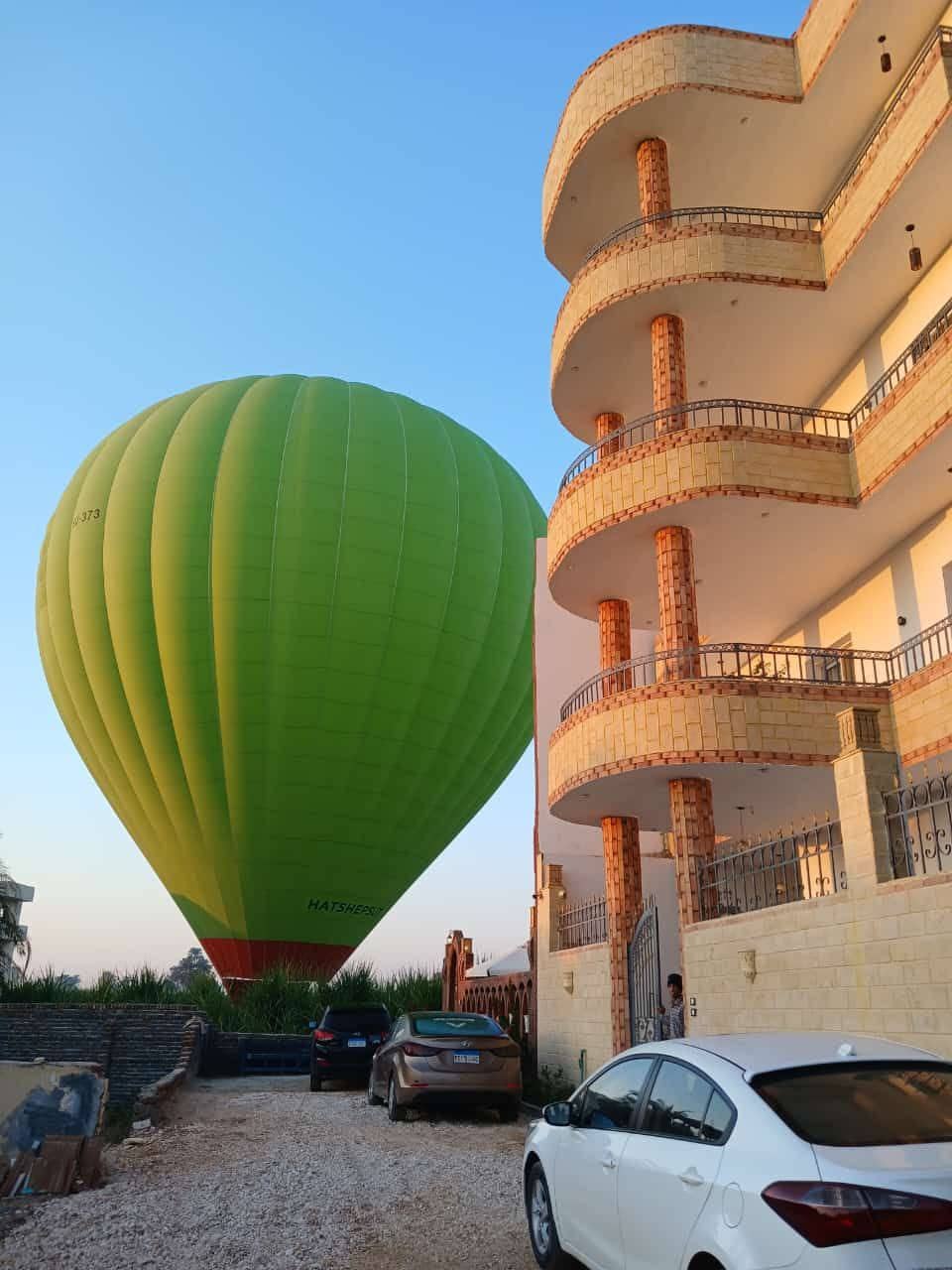 A vibrant green hot air balloon is positioned just beside a multi-level building, casting a large shadow on the ground. Two cars are parked in the driveway, with the balloons providing an engaging focal point against the clear blue sky.