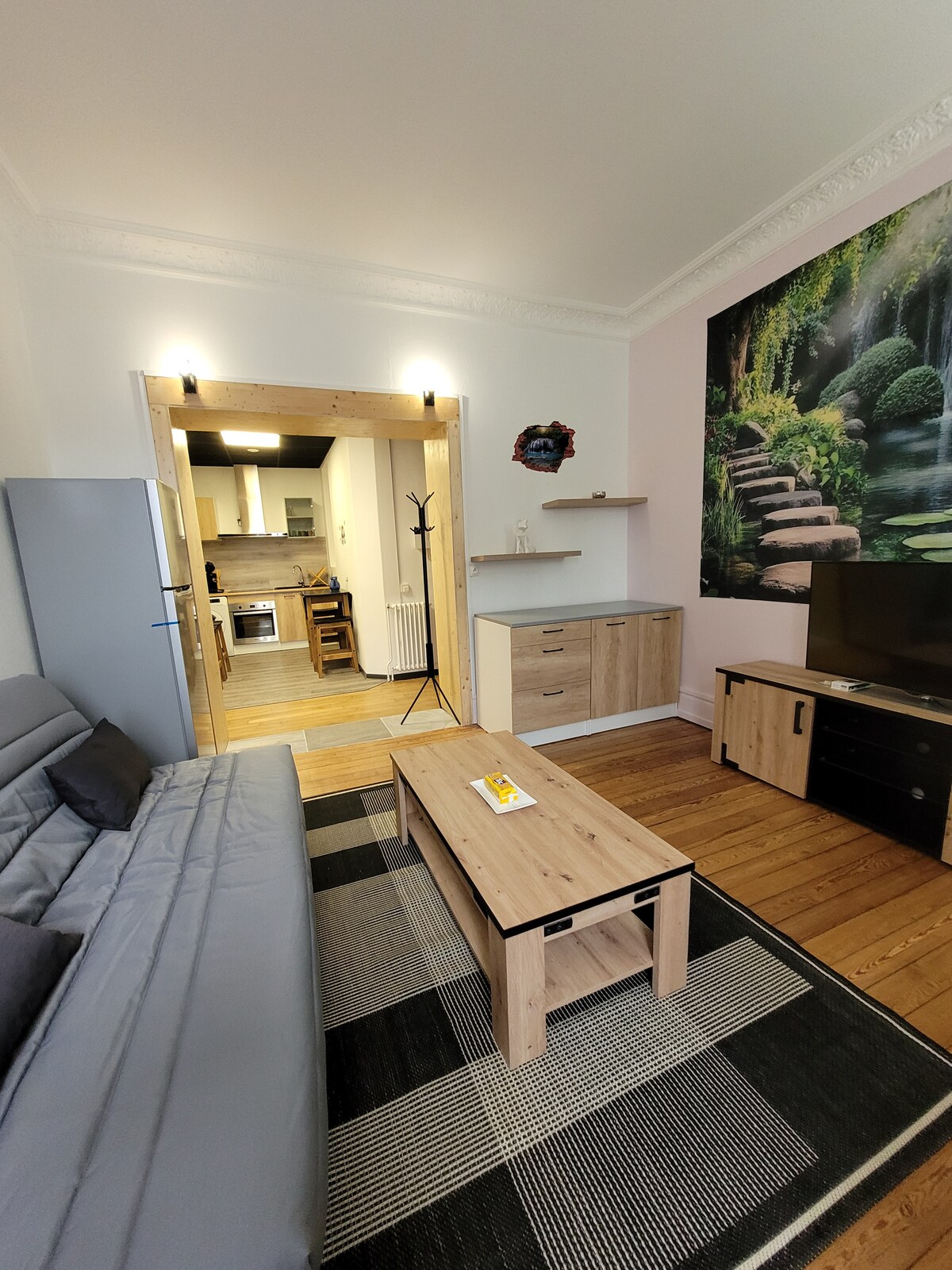 A living area is presented with a gray sofa, wooden coffee table, and a TV stand, creating a comfortable space. A kitchen area is visible in the background, showcasing modern appliances. The wall features a nature-themed mural, contributing to a relaxed ambiance.