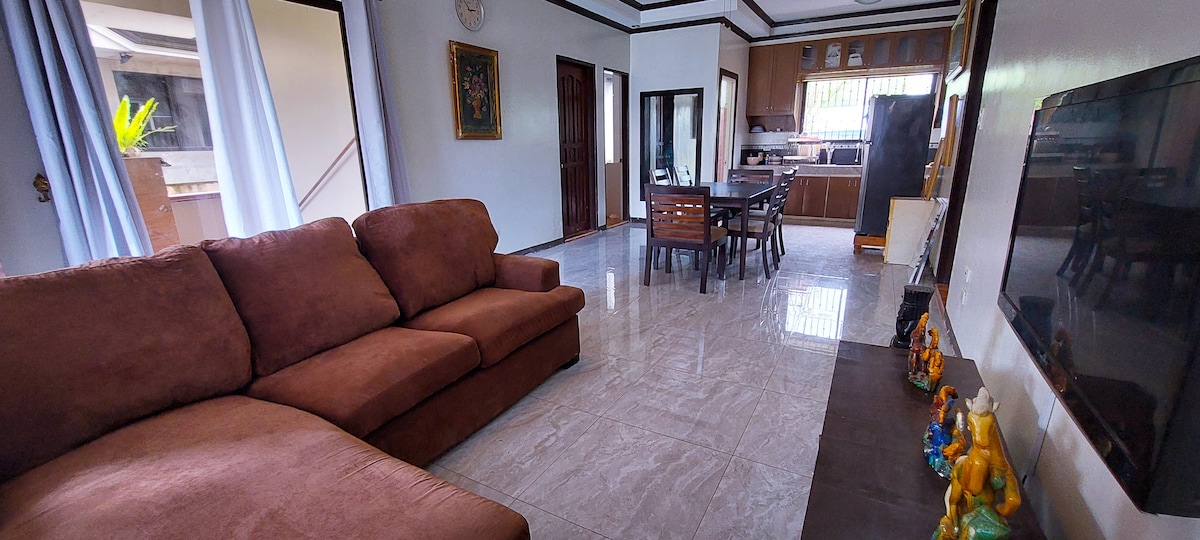 A spacious living area showcases a large brown sofa, complemented by a wooden dining table set for six. The open layout connects to a kitchen area, with natural light coming through large windows and doors. Decorative figurines are displayed on a nearby shelf.