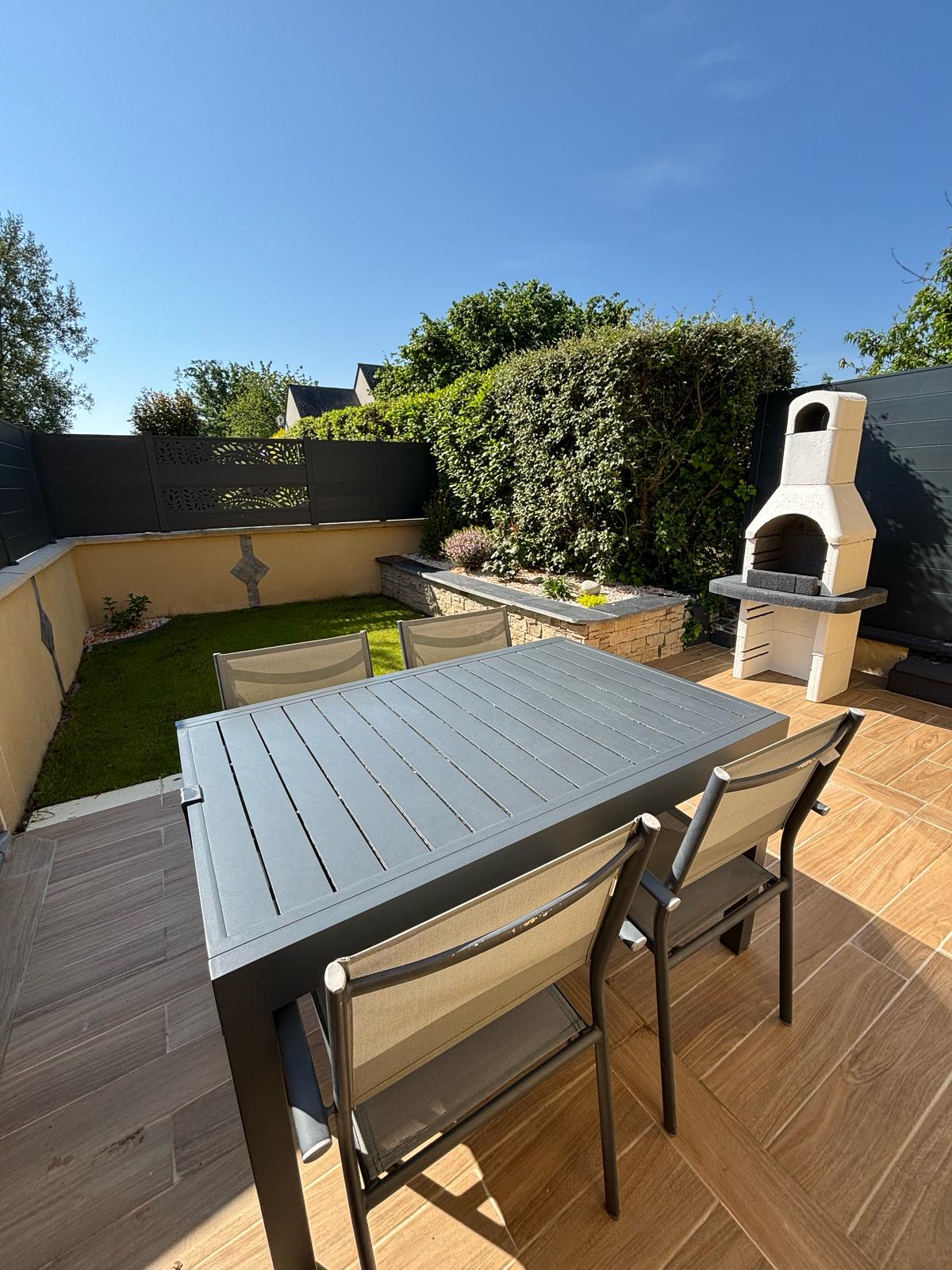 An outdoor terrace is featured, furnished with a dining table and four chairs. A stone barbecue stands nearby, adjacent to a well-maintained garden area. Lush greenery provides a natural backdrop against a clear blue sky.