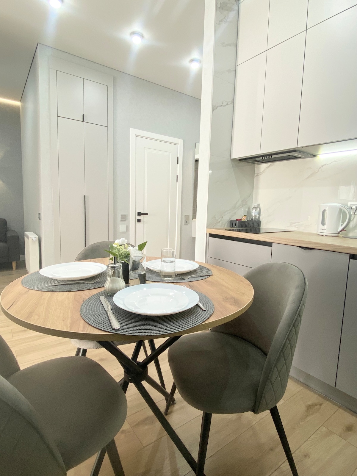 A modern kitchen area is showcased, featuring a round wooden dining table set for four. Stylish upholstered chairs accompany the table. Cabinets with sleek finishes line the walls, and additional appliances are neatly arranged on the countertop, highlighting a functional and inviting cooking space.