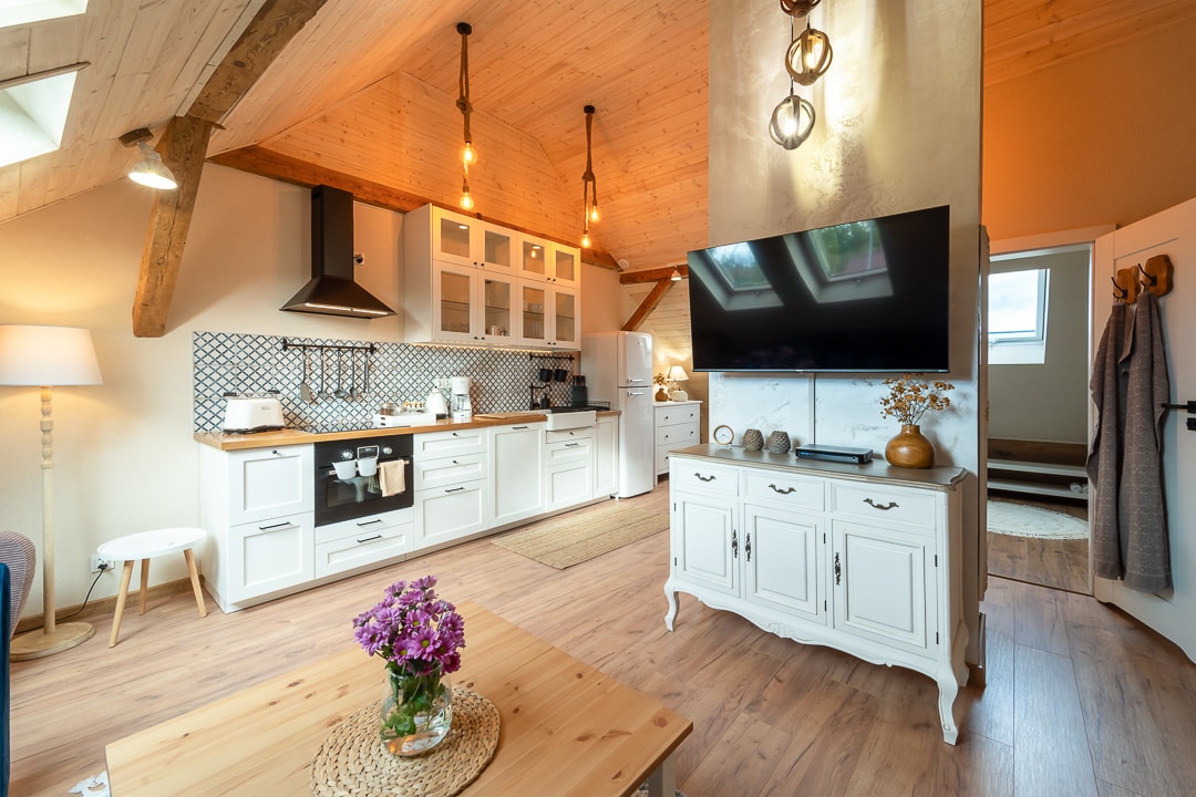 A bright living space features a well-equipped kitchen with white cabinetry and modern appliances. A large screen TV is mounted above a stylish white cabinet. Natural light is invited through skylights and a nearby window, while a small flower arrangement adds a touch of color to the coffee table.