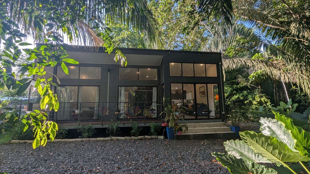The modern villa is surrounded by lush greenery, revealing large full-length windows that offer expansive views of the jungle. A spacious patio leads to the entrance, framed by colorful plants and a gravel pathway that enhances the natural setting.