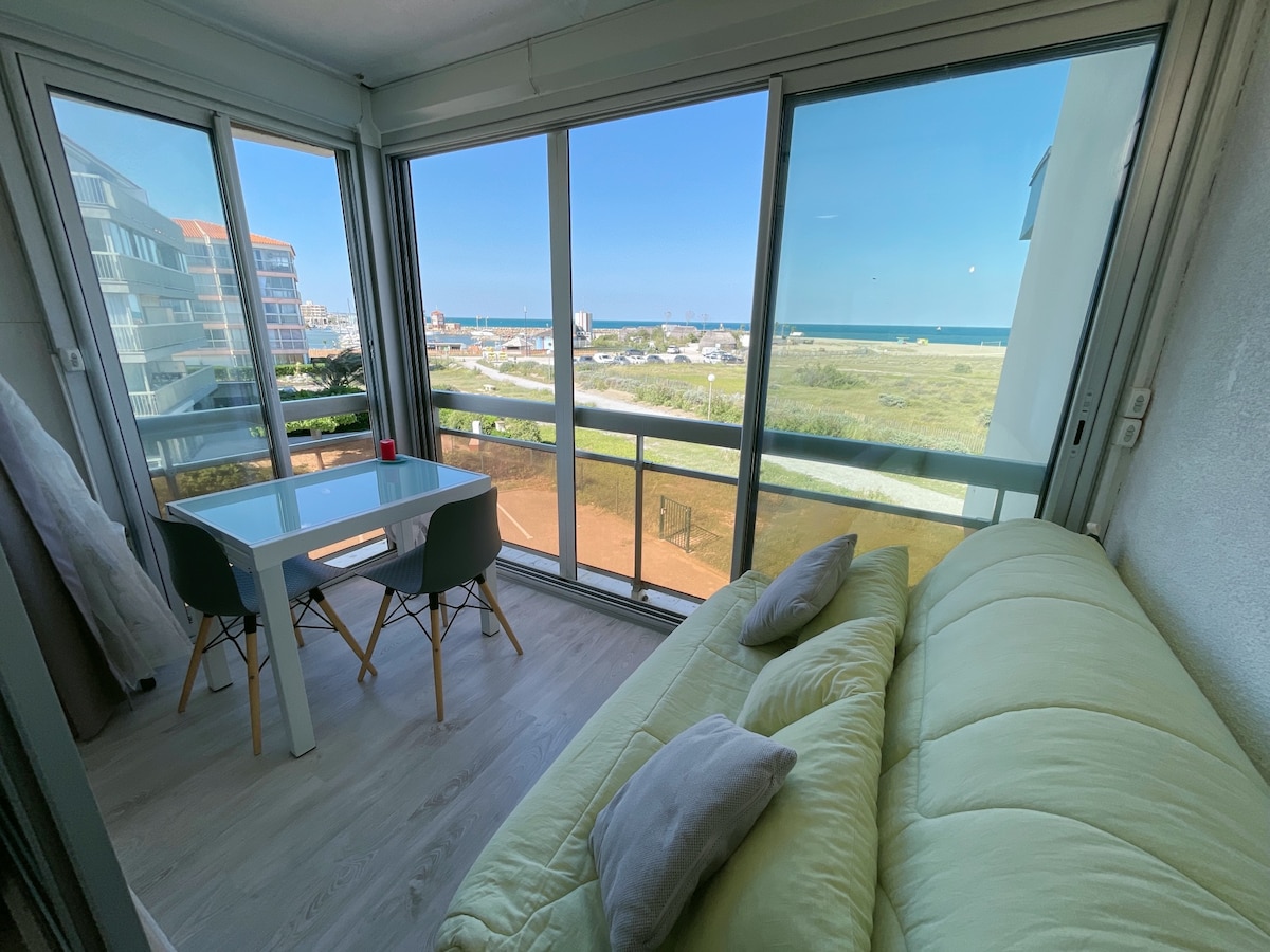A sunlit room features large glass windows providing a view of the Mediterranean and nearby coast. A dining table with chairs occupies one corner, while a light-colored couch with cushions is positioned nearby, creating a comfortable and inviting space.