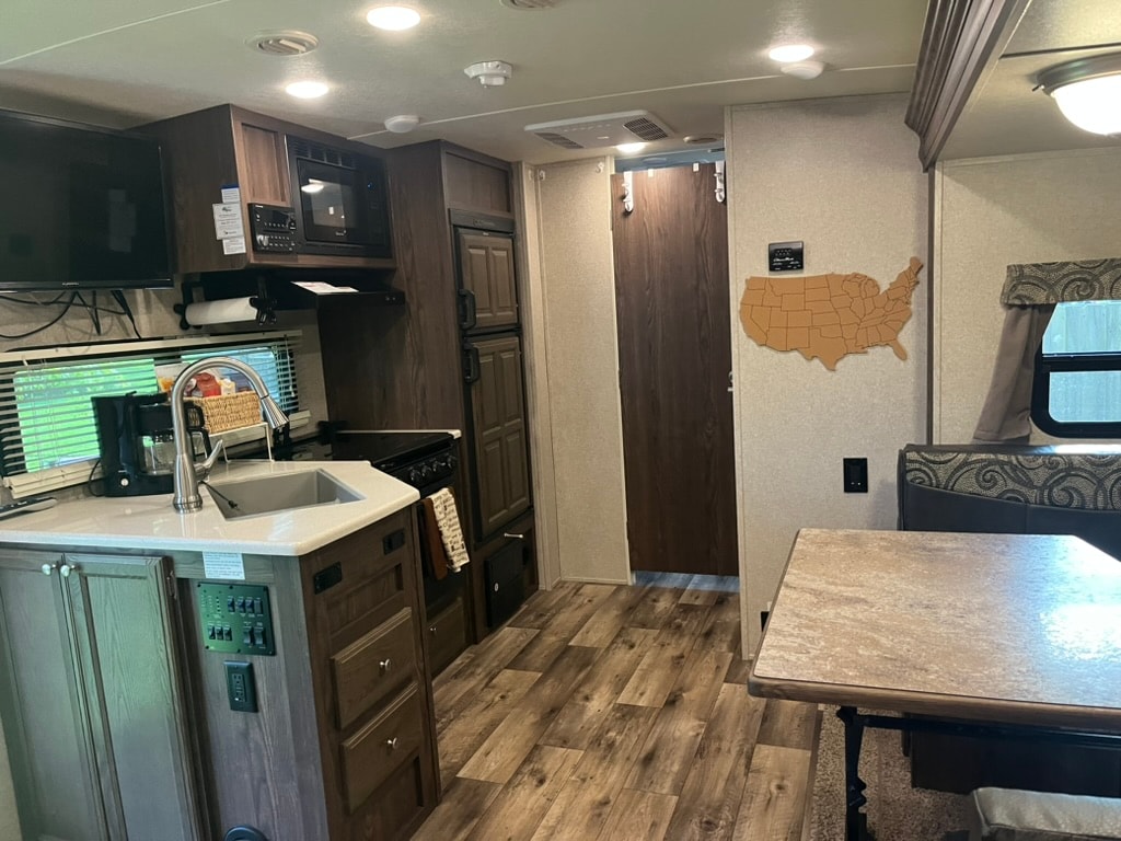 The interior of the travel trailer features a compact kitchen with a sink, stove, microwave, and an ample countertop. A dinette area is visible, accompanied by wood grain flooring. A door leads to the outside, and a map of the United States is mounted on the wall.