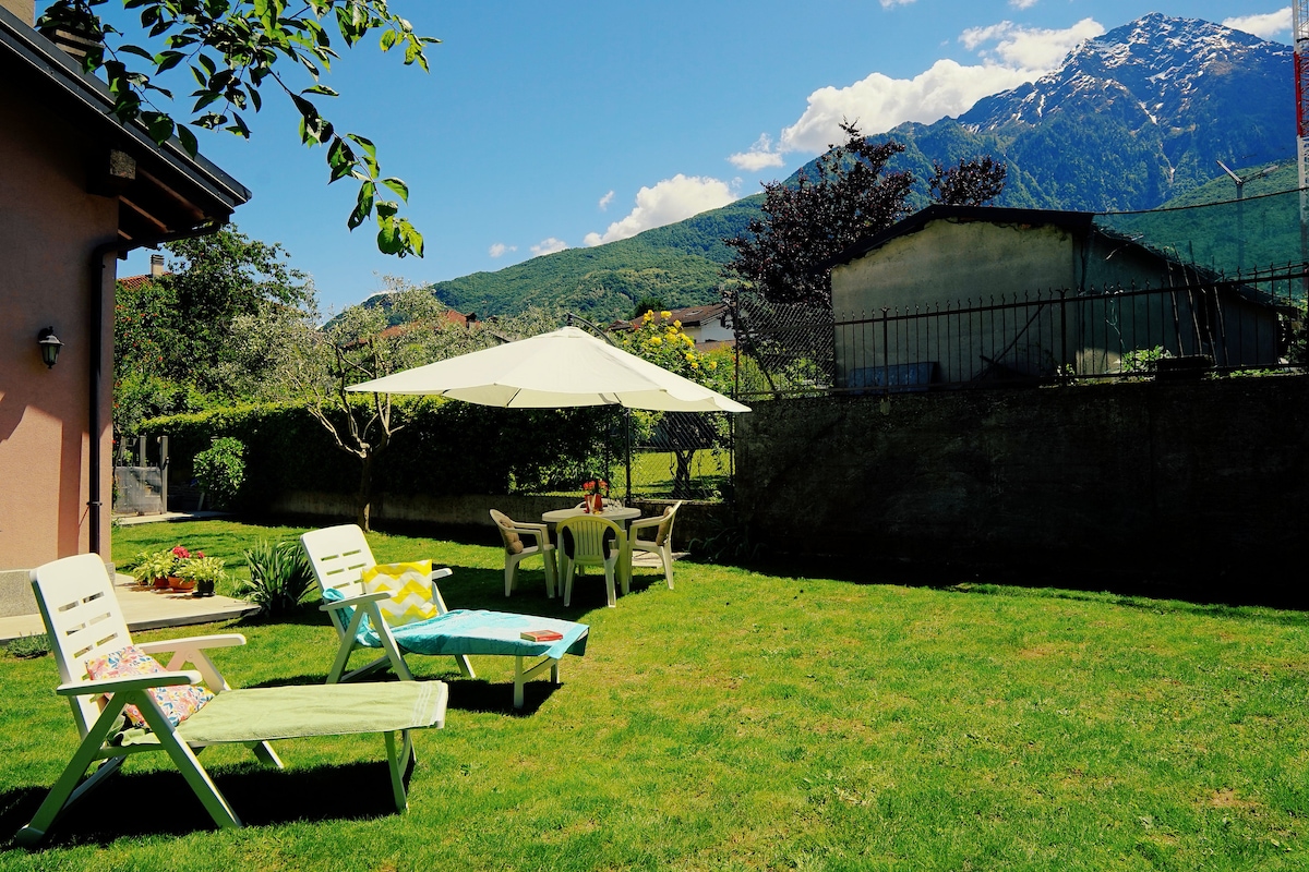 A spacious private garden is presented with a lush green lawn, comfortably furnished with two sun loungers and a dining table under a large umbrella. In the background, distant mountains are visible, enhancing the serene outdoor environment.