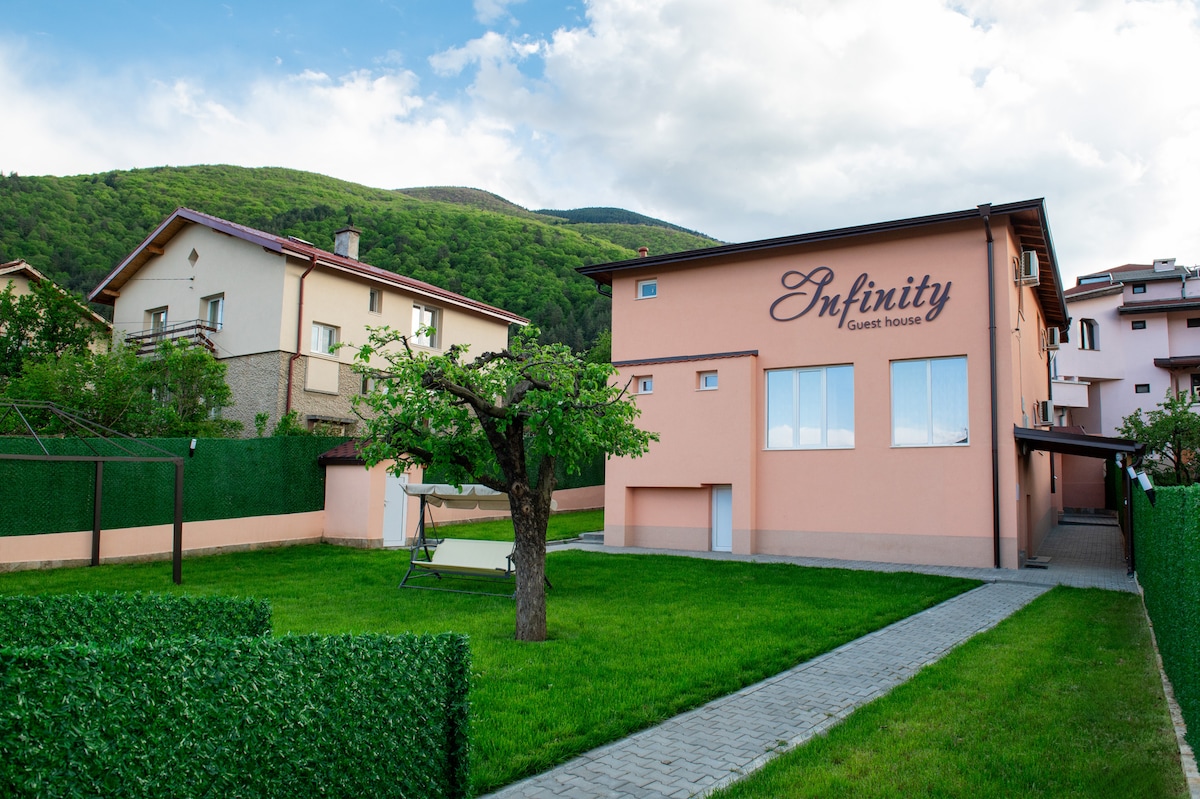 A two-story guest house with a welcoming facade is set against a backdrop of green mountains. The property features a neatly landscaped lawn with a swing and shaded seating area. A walking path is lined with hedges, leading to the entrance of the house.