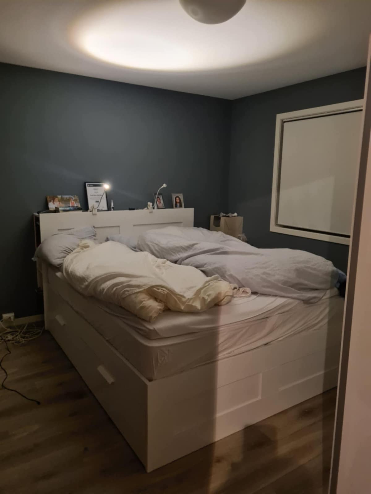 The bedroom features a light-colored bed with a soft, unmade duvet and pillows. A bedside table with a small lamp is positioned beside the bed. Dark blue walls provide a calming backdrop, and a framed window is visible, allowing natural light to soften the space.