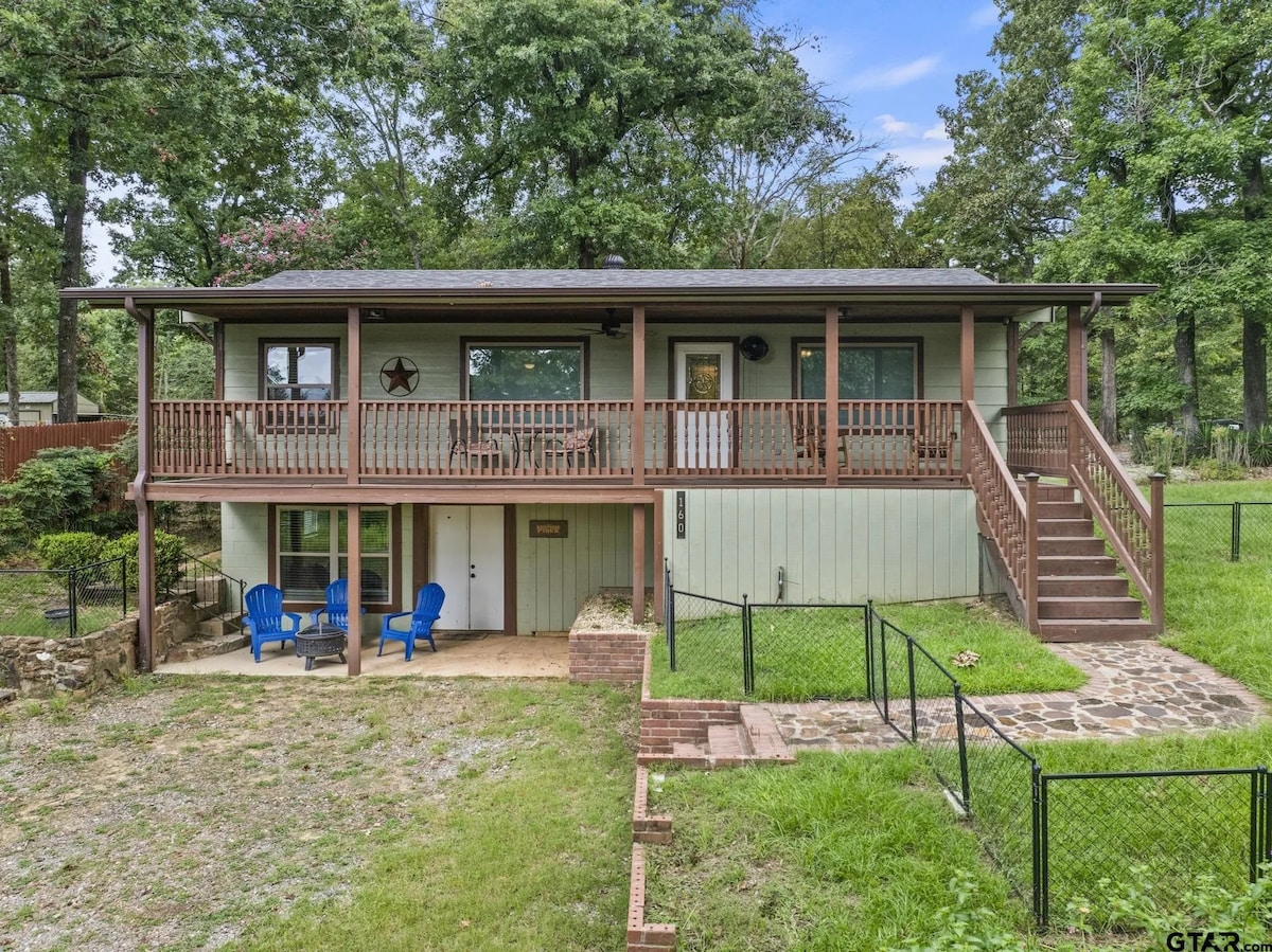 The exterior of a two-story house is showcased, featuring a front porch with chairs. A grassy yard surrounds the structure, and a stone pathway leads to the entrance. The facade incorporates a mix of wood and green siding, complemented by natural landscaping.