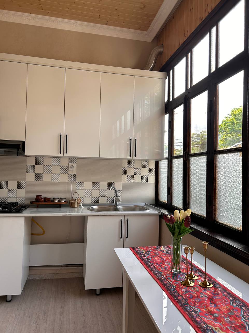 A well-organized kitchen is presented with white cabinetry and a modern countertop. Natural light filters through large windows, illuminating the space. A small dining table is adorned with a patterned cloth and a vase of tulips, while decoratively arranged candle holders add subtle elegance.