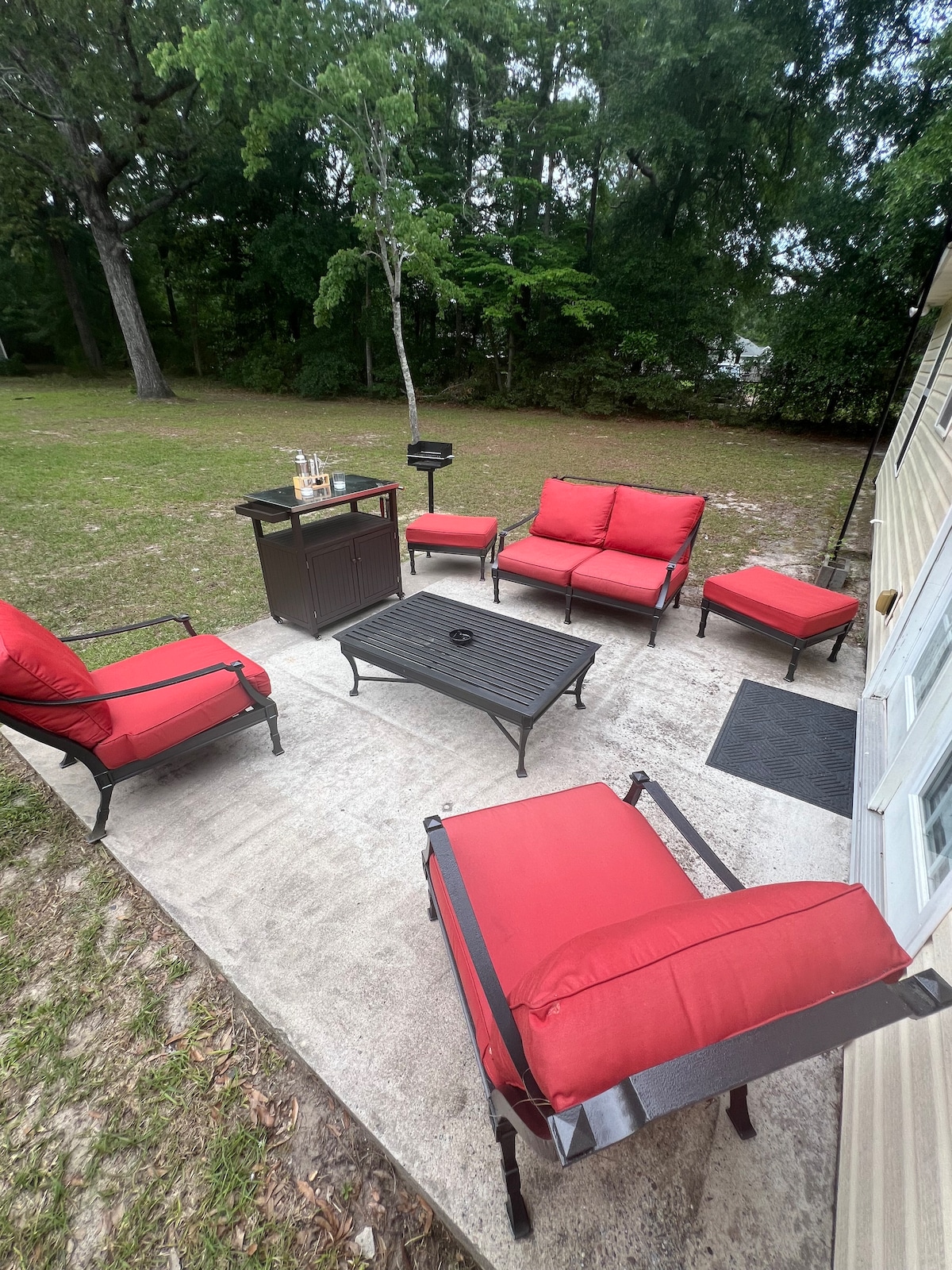 An outdoor patio area features red cushioned seating arranged around a black metal coffee table. A small grill is positioned nearby on a concrete surface, surrounded by a grassy space with trees in the background, providing a relaxed setting for gatherings.