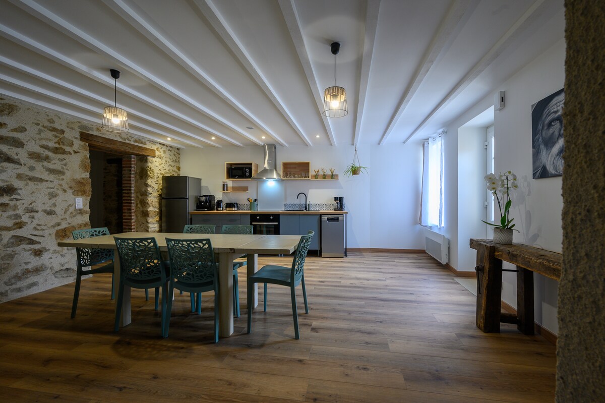 An open dining area is presented with a large wooden table surrounded by six blue chairs. Modern kitchen appliances and cabinetry are visible in the background, while natural light filters through a window, highlighting the rustic stone wall and wooden beams on the ceiling.