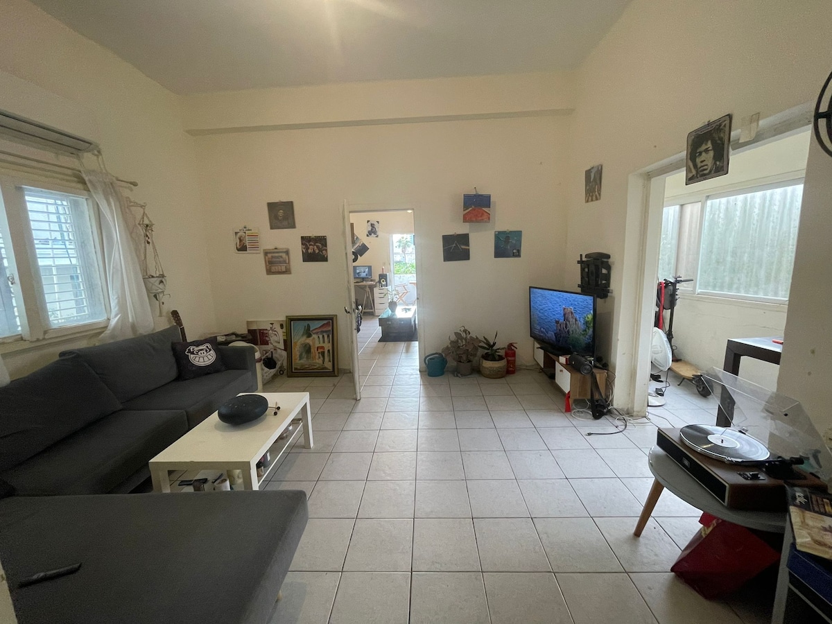 The living room features a spacious gray sofa and a coffee table. Light-colored tiles cover the floor. A television sits on the side, and various artworks decorate the walls. Sunlight enters through multiple windows, creating a bright and open atmosphere.