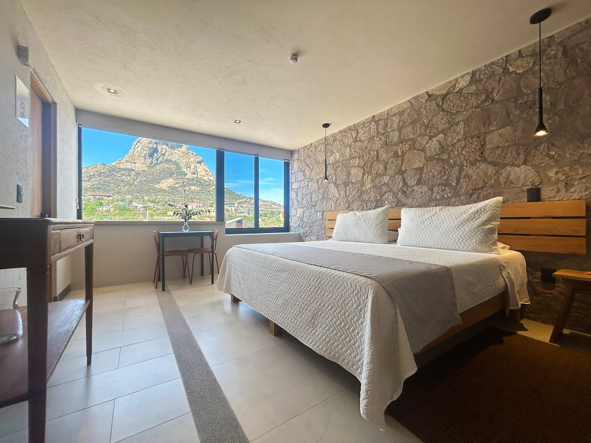 A spacious sleeping area features two beds, complemented by stone wall accents. Large windows offer a view of Peña de Bernal. A small desk and chair are positioned near the window, with soft natural light illuminating the room.