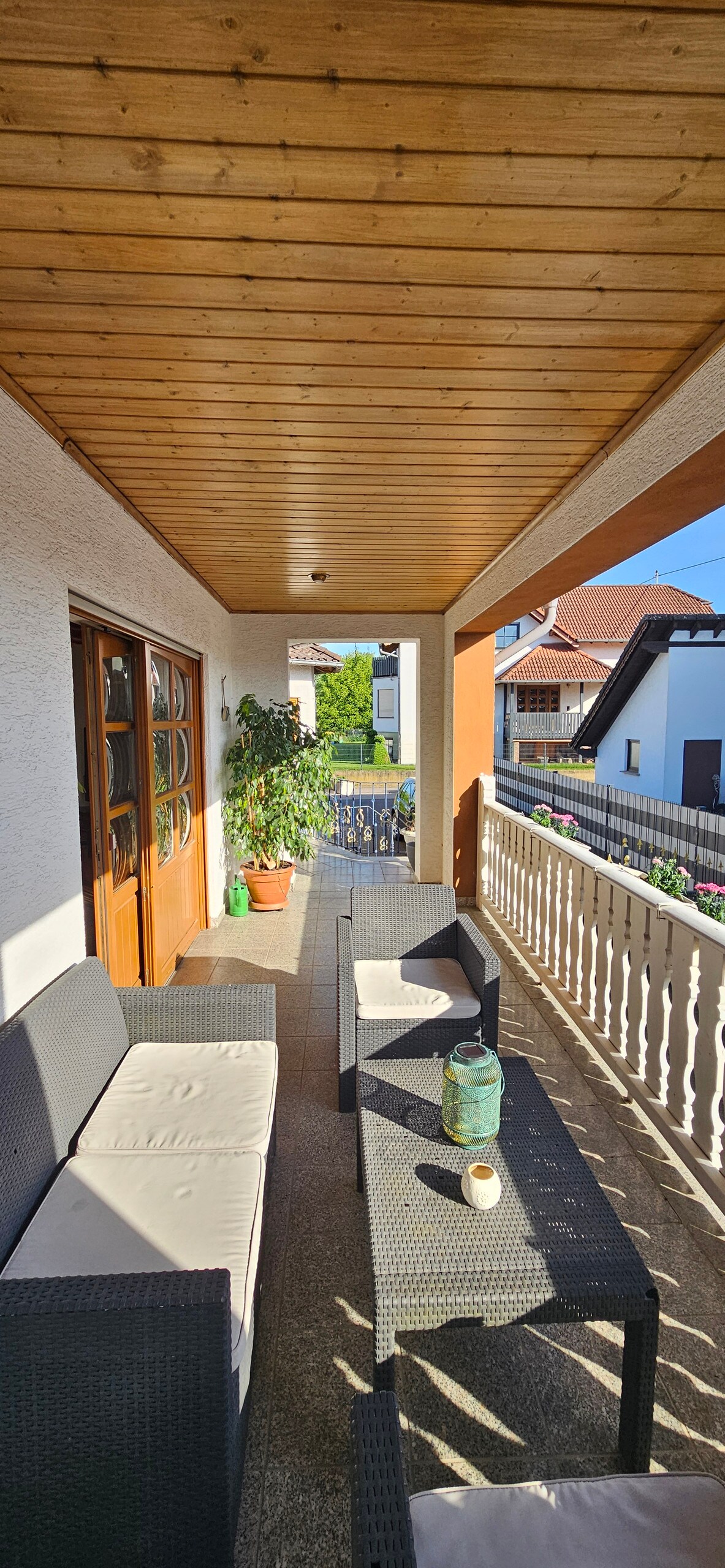 A covered balcony features a wooden ceiling and is furnished with a light-colored seating arrangement. Two armchairs and a sofa are positioned around a small table. A potted plant adds a touch of greenery, while natural light illuminates the welcoming space.