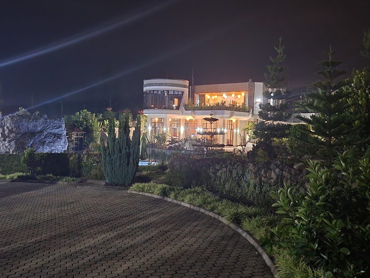 The resort's exterior is illuminated at night, revealing a modern structure surrounded by lush vegetation. Pathways made of stone lead to the entrance, enhancing the overall tranquility of the setting. Soft lighting highlights the building's design and creates a cozy environment.