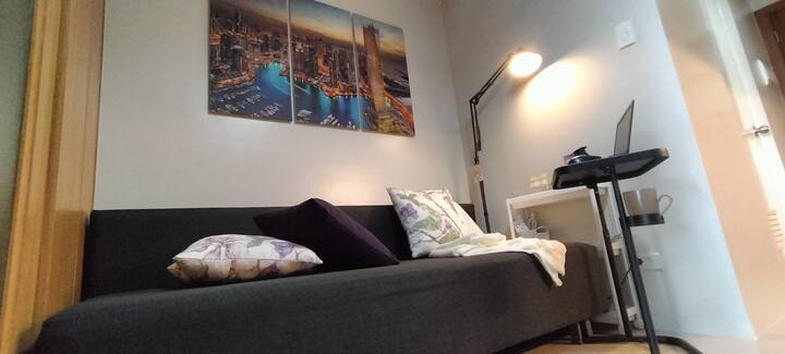 Un espacio acogedor y moderno con un sofá cama negro con lujosas almohadas florales, un espacio de trabajo funcional con una laptop y una lámpara de escritorio y vibrantes obras de arte del paisaje urbano. El espacio luminoso y limpio es perfecto para relajarse y trabajar.