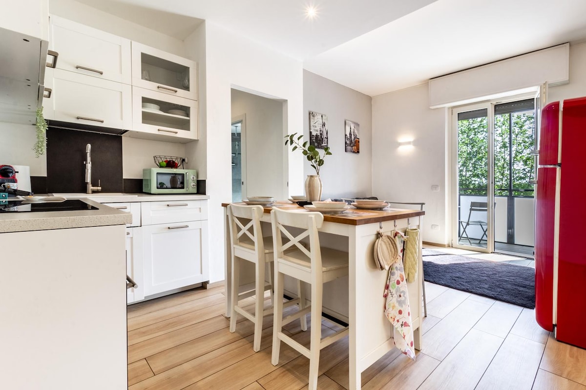 A modern kitchen is showcased, featuring white cabinetry and essential appliances, including a microwave and a red refrigerator. A central island with high stools provides a dining area. A glass door leads to a small balcony with outdoor seating, inviting natural light into the space.