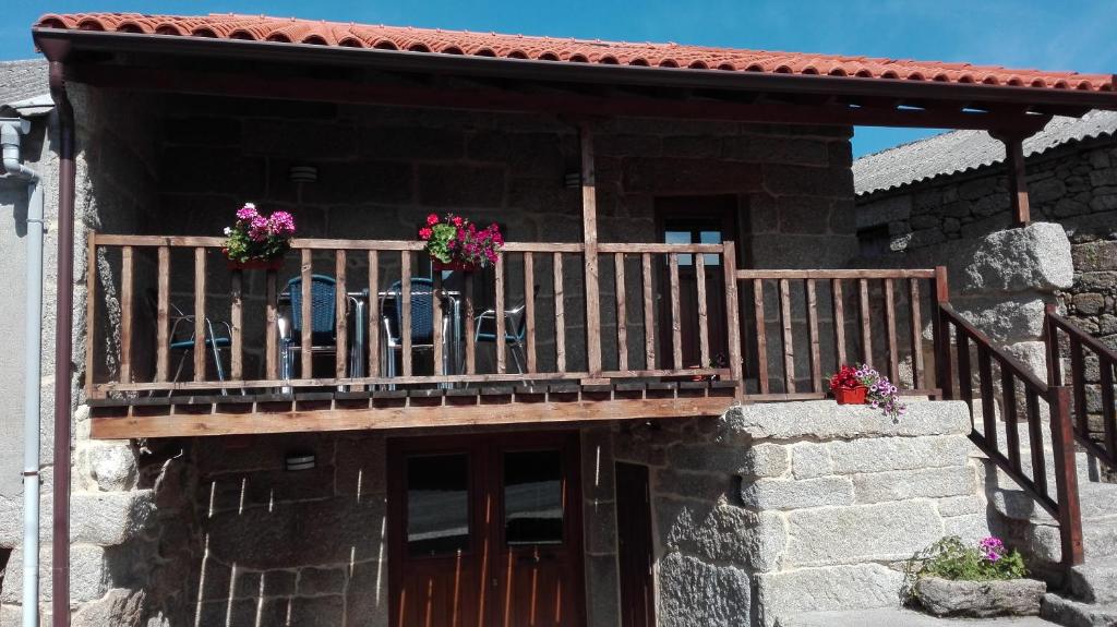 A rustic stone house features a wooden balcony adorned with colorful flowers. Two black chairs and a small table are positioned on the balcony, inviting guests to relax outdoors. The entrance is framed by sturdy wooden doors, and a clear blue sky is visible above.